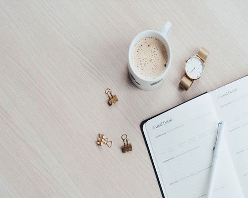 A flatlay of an organized workspace showing a ceramic mug, a water flask, and an analog notebook on a light surface